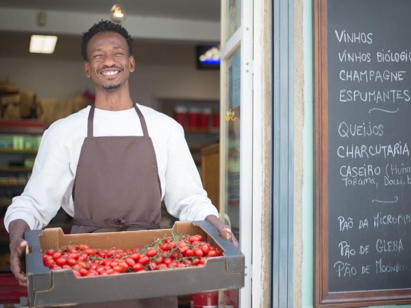 Smiling vendor standing outside a Portuguese store holding a box of ripe tomatoes. Perfect for grocery or food themes.